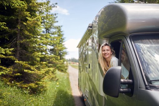 Eine Frau lehnt sich lächelnd aus dem Fenster eines Wohnmobils auf einer schmalen Landstraße inmitten grüner Natur – Symbol für Freiheit, Campingurlaub und sicheres Reisen.
