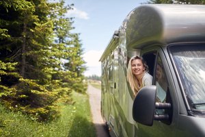 Eine Frau lehnt sich lächelnd aus dem Fenster eines Wohnmobils auf einer schmalen Landstraße inmitten grüner Natur – Symbol für Freiheit, Campingurlaub und sicheres Reisen.