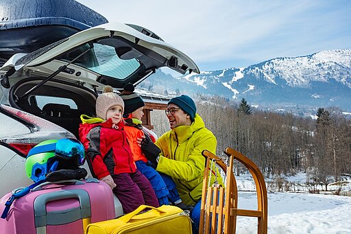 Eine Familie bereitet sich auf einen Winterurlaub vor. Im Vordergrund sind bunte Koffer und Taschen zu sehen, während ein Erwachsener einem Kind mit roter Jacke und Mütze hilft, sich warm anzuziehen. Im Hintergrund erstreckt sich eine schneebedeckte Berglandschaft unter einem strahlend blauen Himmel, was die winterliche Atmosphäre unterstreicht.