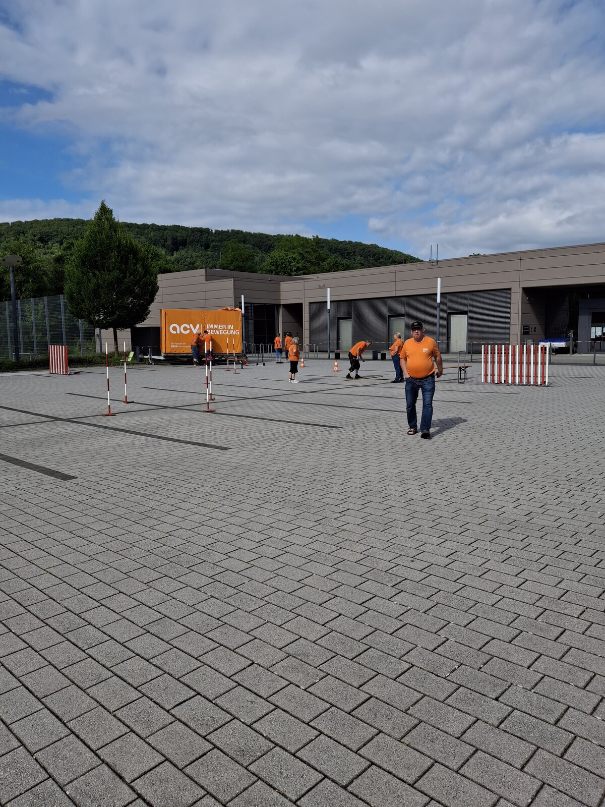 Vier Personen in orangefarbenen T-Shirts bauen auf einem gepflasterten Außenbereich in der Nähe eines modernen Gebäudes einen Parcours aus Stangen und Absperrungen auf; im Hintergrund sind unter einem teilweise bewölkten Himmel ein großer orangefarbener Lkw und grüne Bäume zu sehen.