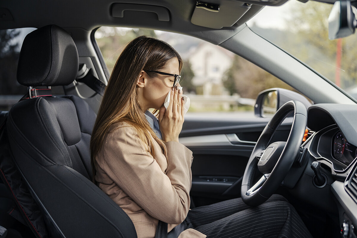 Eine Frau sitzt auf dem Fahrersitz eines Autos, trägt eine Brille und bedeckt ihre Nase und ihren Mund mit einem Taschentuch, als ob sie niesen oder eine Allergie haben würde. Sie ist mit einem beigen Mantel bekleidet und hat braunes Haar.