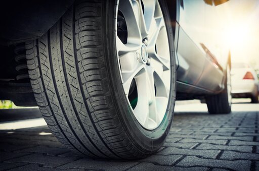 Nahaufnahme eines Autoreifens auf einer Felge aus einer Silberlegierung, der auf einer gepflasterten Fläche steht, während im Hintergrund Sonnenlicht scheint und rechts ein weiteres Auto leicht zu erkennen ist.