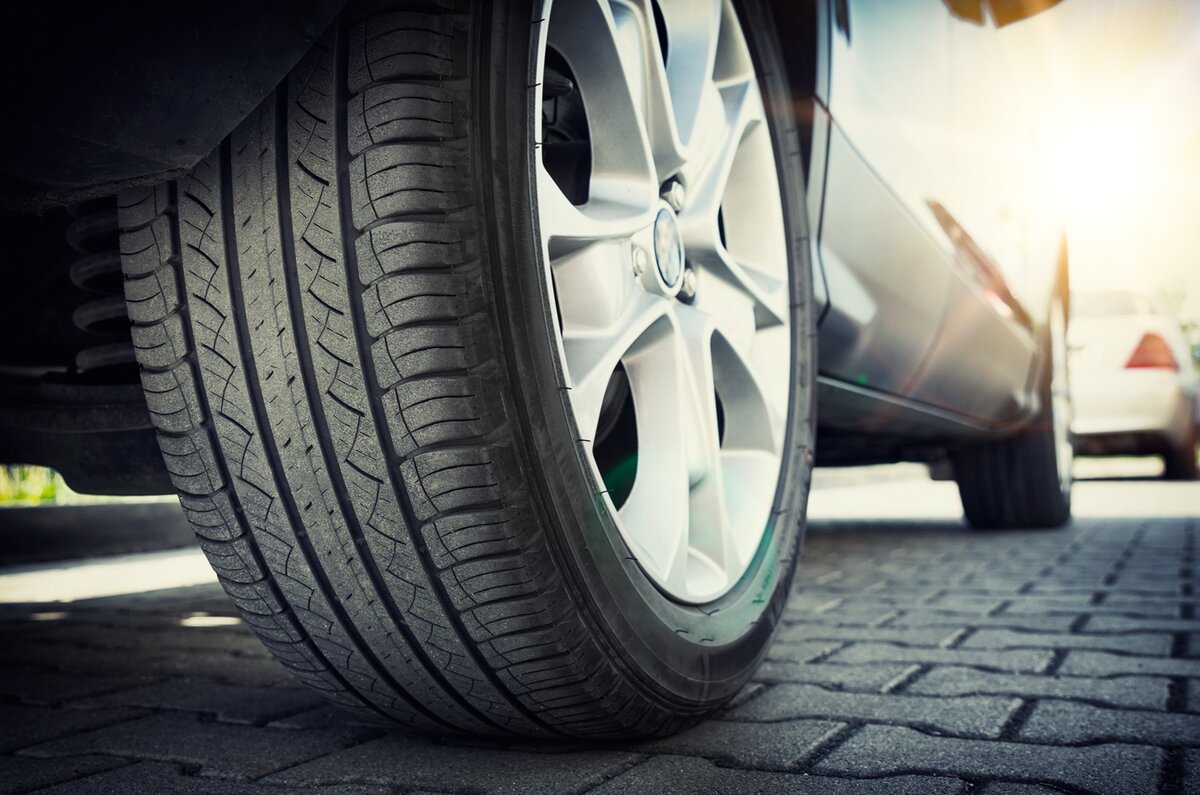 Nahaufnahme eines Autoreifens auf einer Felge aus einer Silberlegierung, der auf einer gepflasterten Fläche steht, während im Hintergrund Sonnenlicht scheint und rechts ein weiteres Auto leicht zu erkennen ist.