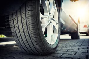 Nahaufnahme eines Autoreifens auf einer Felge aus einer Silberlegierung, der auf einer gepflasterten Fläche steht, während im Hintergrund Sonnenlicht scheint und rechts ein weiteres Auto leicht zu erkennen ist.