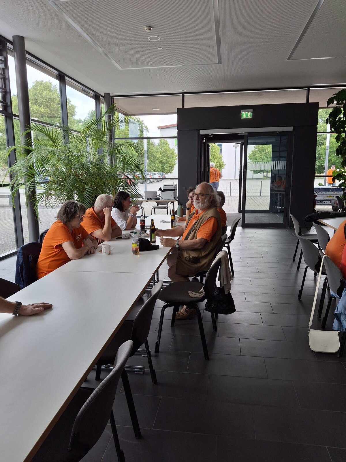 Eine Gruppe älterer Menschen in orangefarbenen T-Shirts sitzt um einen Tisch herum in einem modernen, hellen Raum mit großen Fenstern, Grünpflanzen und Getränken auf dem Tisch. Einige unterhalten sich, und einer schaut in die Kamera.
