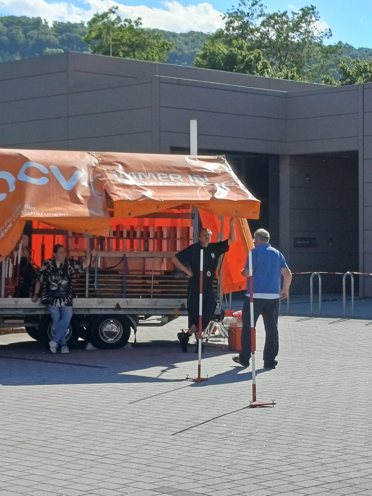 Drei Personen stehen in einem sonnigen Außenbereich neben einem Anhänger mit einem orangefarbenen Vordach. Im Vordergrund stehen zwei Pfosten, im Hintergrund sind ein modernes Gebäude und grüne Bäume zu sehen.