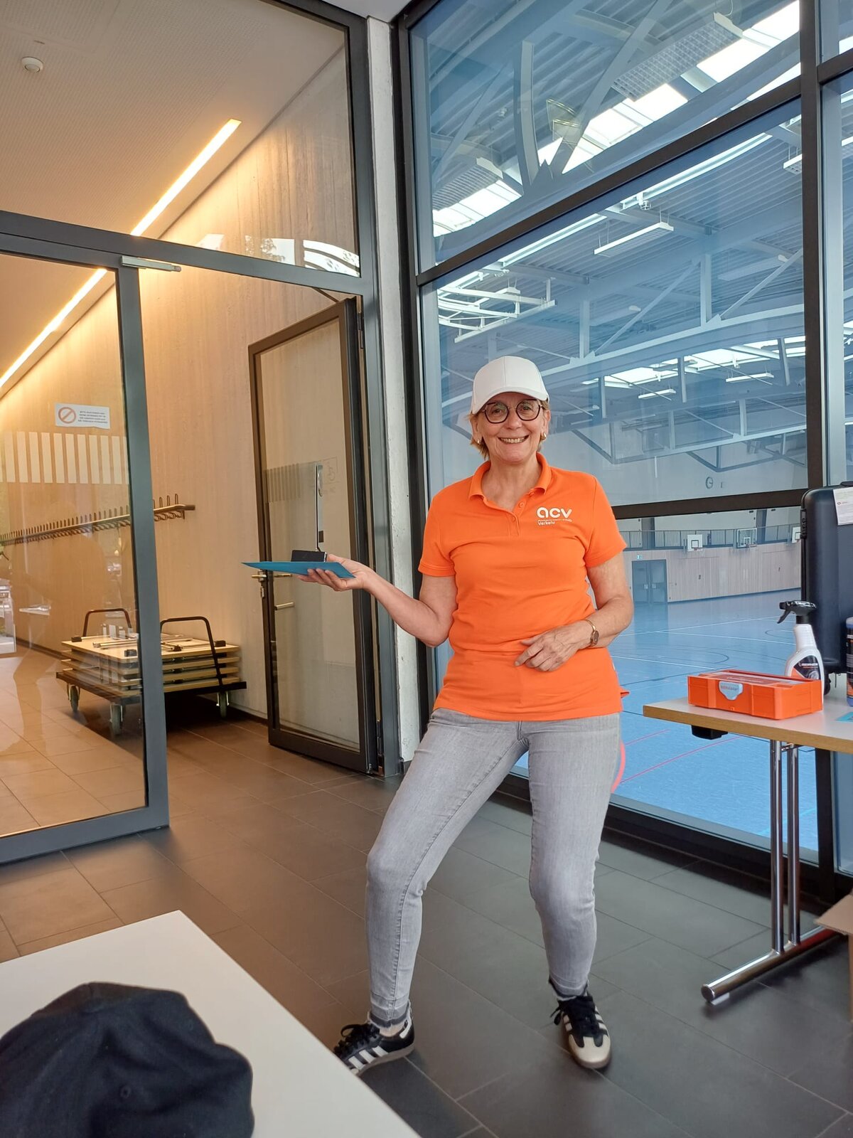Eine Frau in einem orangefarbenen Polohemd, grauen Jeans und einer weißen Baseballkappe, die lächelt und eine blaue Mappe in der Hand hält, in einem Gebäude mit großen Fenstern.