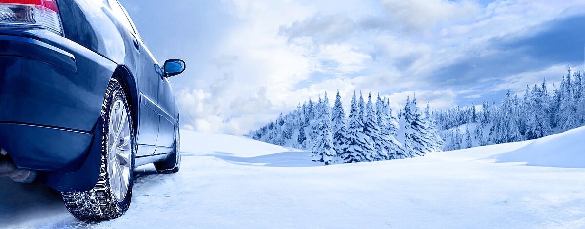 Auto mit Winterreifen fährt auf verschneiter Straße durch winterliche Landschaft mit Tannen und Schnee.