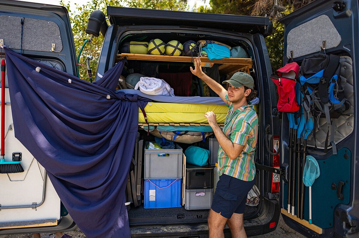 Eine Person steht neben einem offenen Lieferwagen, der mit Campingausrüstung vollgepackt ist, und ordnet die Gegenstände unter einem Hochbett. In den Türen des Lieferwagens sind Rucksäcke und andere Ausrüstungsgegenstände verstaut, und an der Seite hängt ein blauer Stoff, im Hintergrund sind Bäume zu sehen.