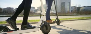 Zwei Personen in Geschäftskleidung fahren mit Elektrorollern auf einem städtischen Weg, im Hintergrund sind moderne Bürogebäude und Grünflächen zu sehen. Das Bild wurde aus einem niedrigen Blickwinkel aufgenommen, wobei der Fokus auf den Rollern und den Beinen der Personen liegt.