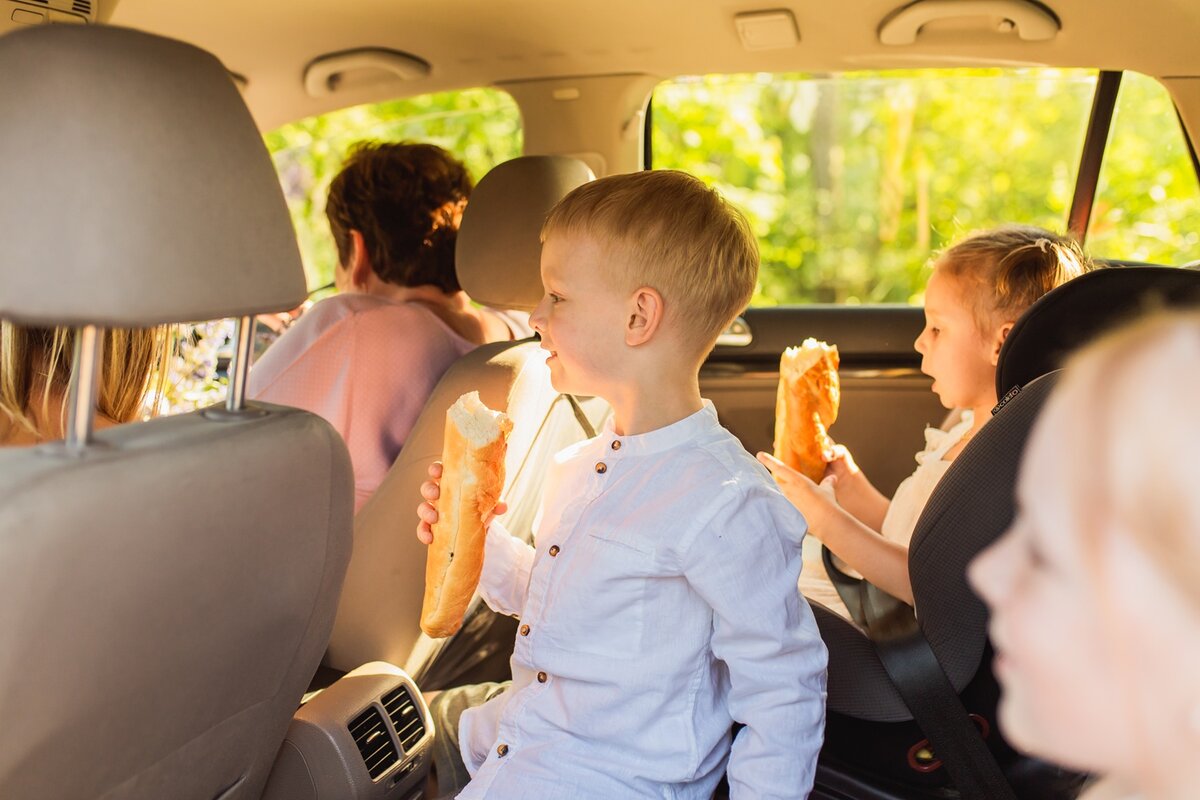Drei kleine Kinder sitzen auf der Rückbank eines Autos und essen Baguette, während ein Erwachsener auf dem Vordersitz sitzt.