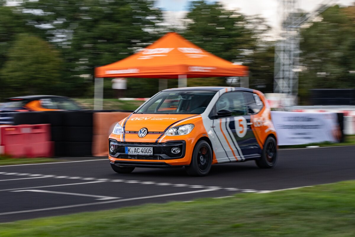 Ein orange-weißer Rennwagen mit der Nummer 5 rast über eine Rennstrecke. Im Hintergrund sind ein orangefarbenes Zelt und Bäume zu sehen.