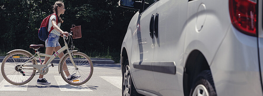 Ein Kind schiebt ihr Fahrrad über ein Zebrastreifen und ein weißes Auto wartet