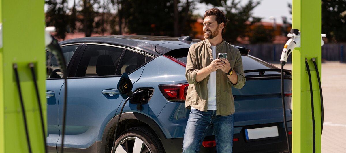 Ein Mann lehnt sich an ein blaues Elektroauto, während er es an einer öffentlichen Ladestation auflädt, hält sein Handy in der Hand und lächelt. Das Ladekabel ist an das Auto angeschlossen, und die Szene spielt draußen auf einem Parkplatz.