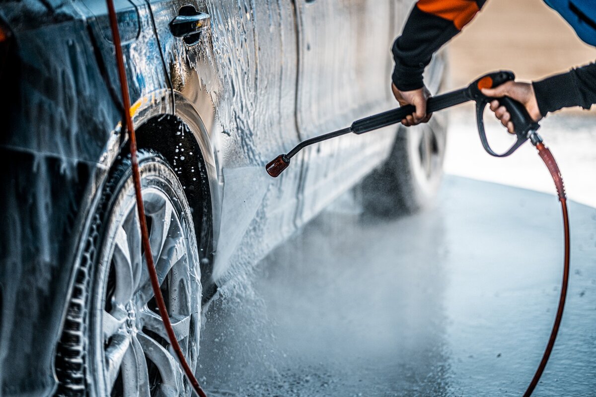 Eine Person wäscht ein mit Seife bedecktes Auto mit einem Hochdruckschlauch, wobei sie sich auf die Reinigung der Räder und des Unterbodens konzentriert.