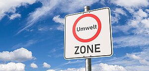 Verkehrsschild Umweltzone mit rotem Rand und Aufschrift Umwelt, darunter Zusatzschild Zone, aufgenommen vor blauem Himmel.