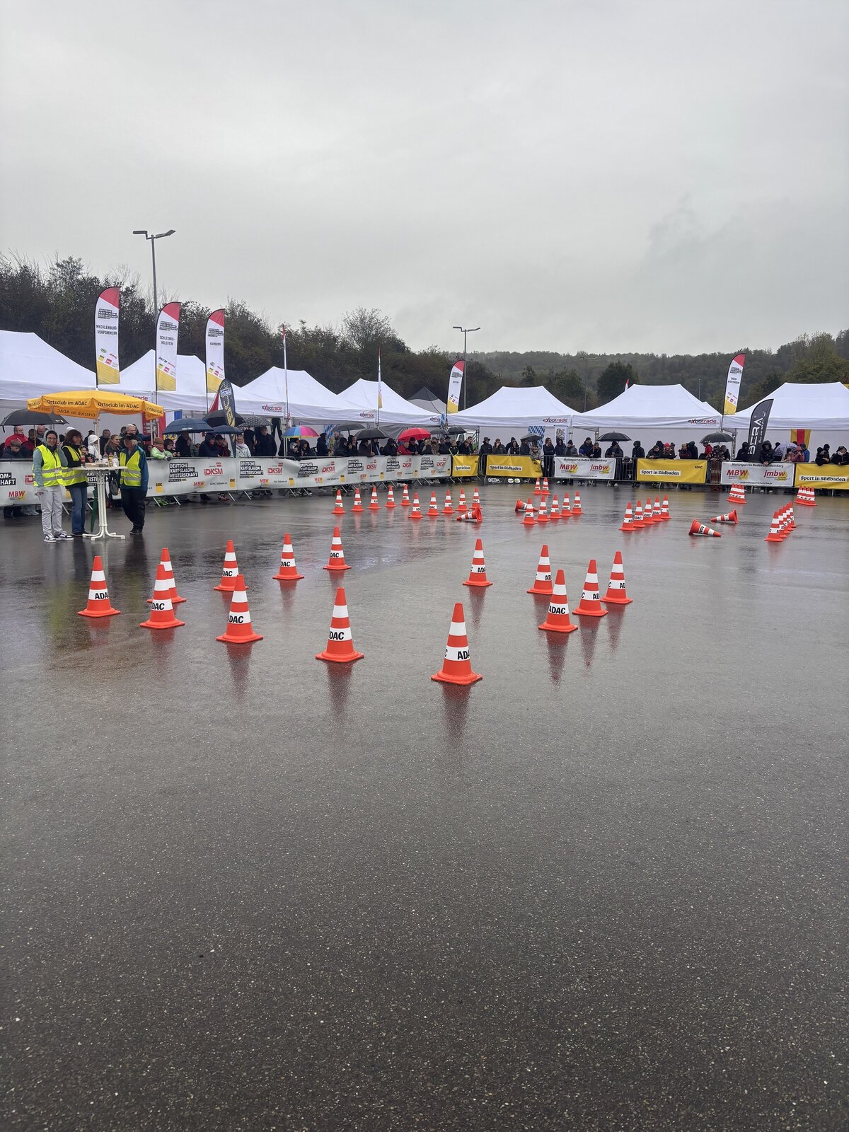 Eine Strecke, die auf einem nassen Asphaltplatz durch zahlreiche orangefarbene Verkehrskegel markiert ist, im Hintergrund sind weiße Zelte und Zuschauer zu sehen.