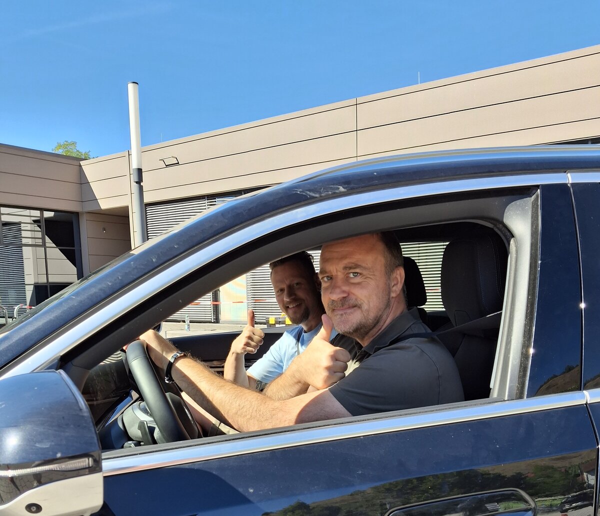 Zwei Männer sitzen in einem Auto, beide strecken den Daumen hoch und lächeln. Das Auto steht an einem sonnigen Tag vor einem modernen Gebäude.