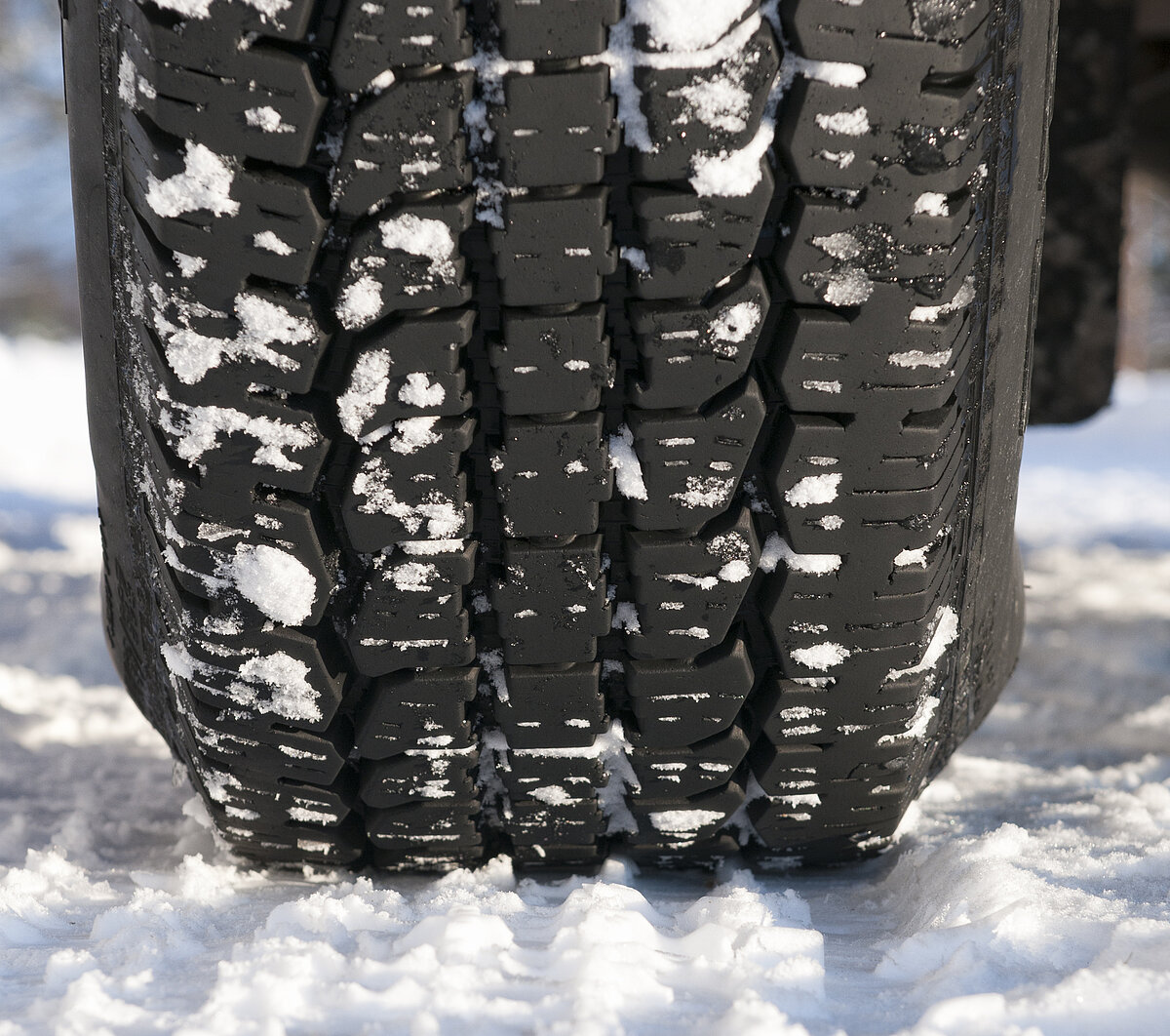 Nahaufnahme eines Winterreifens mit Schneeresten im Profil auf verschneiter Fahrbahn.