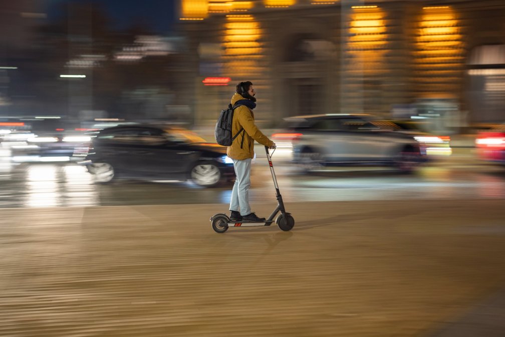 Person fährt nachts mit einem E-Scooter durch den Stadtverkehr – umgeben von Autos und beleuchteten Gebäuden.