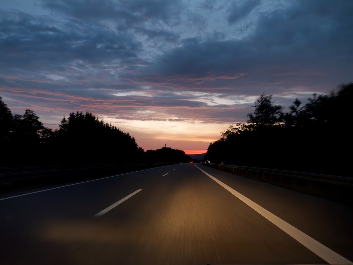 Ein Blick aus einem Auto, das in der Abenddämmerung auf einer leeren Autobahn fährt, mit Bäumen zu beiden Seiten und einem farbenprächtigen Himmel, der von Blau über Orange bis hin zu Rosa wechselt, während die Sonne am Horizont untergeht.