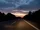 Ein Blick aus einem Auto, das in der Abenddämmerung auf einer leeren Autobahn fährt, mit Bäumen zu beiden Seiten und einem farbenprächtigen Himmel, der von Blau über Orange bis hin zu Rosa wechselt, während die Sonne am Horizont untergeht.