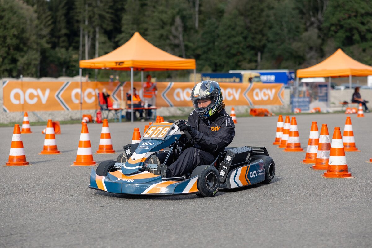 Eine Person, die einen Helm und einen Rennanzug trägt, fährt mit einem blau-orangefarbenen Go-Kart mit der Startnummer 508 durch einen mit orangefarbenen Verkehrskegeln markierten Parcours.