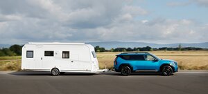 Ein blauer Geländewagen zieht einen großen weißen Wohnwagen auf einer asphaltierten Straße, mit Feldern und fernen Hügeln unter einem teilweise bewölkten Himmel im Hintergrund.
