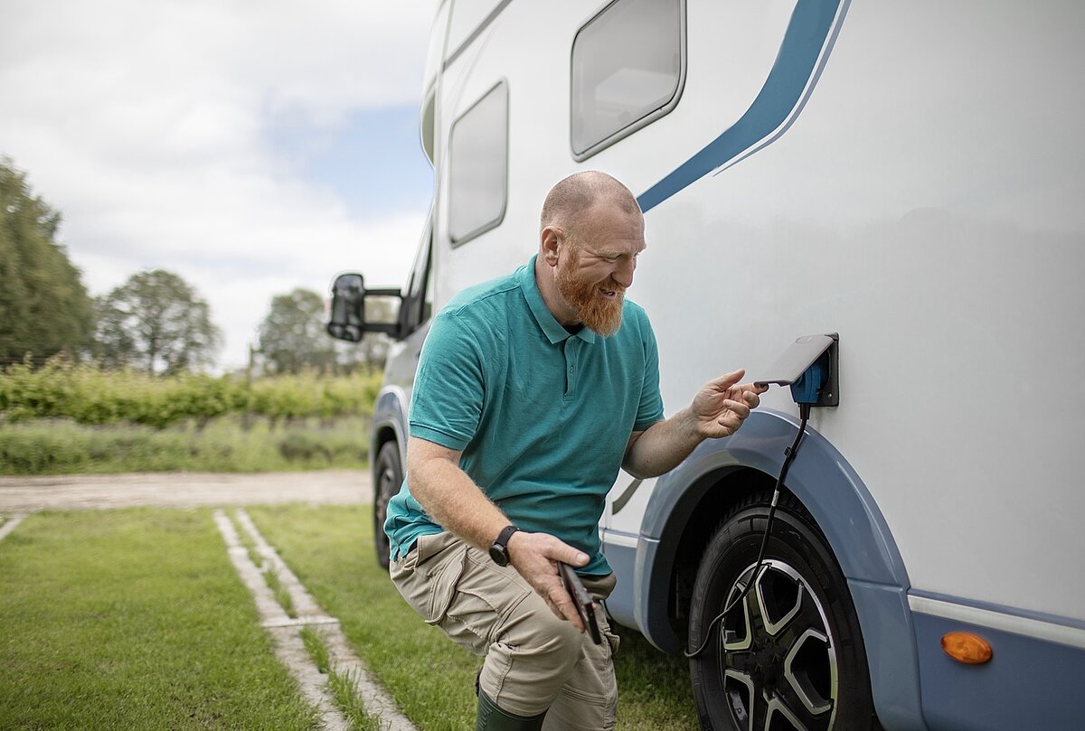 Ein Mann kniet neben einem weißen Wohnmobil und lächelt, während er ein Ladekabel an das Fahrzeug anschließt oder abtrennt, mit grünem Gras und Bäumen im Hintergrund.