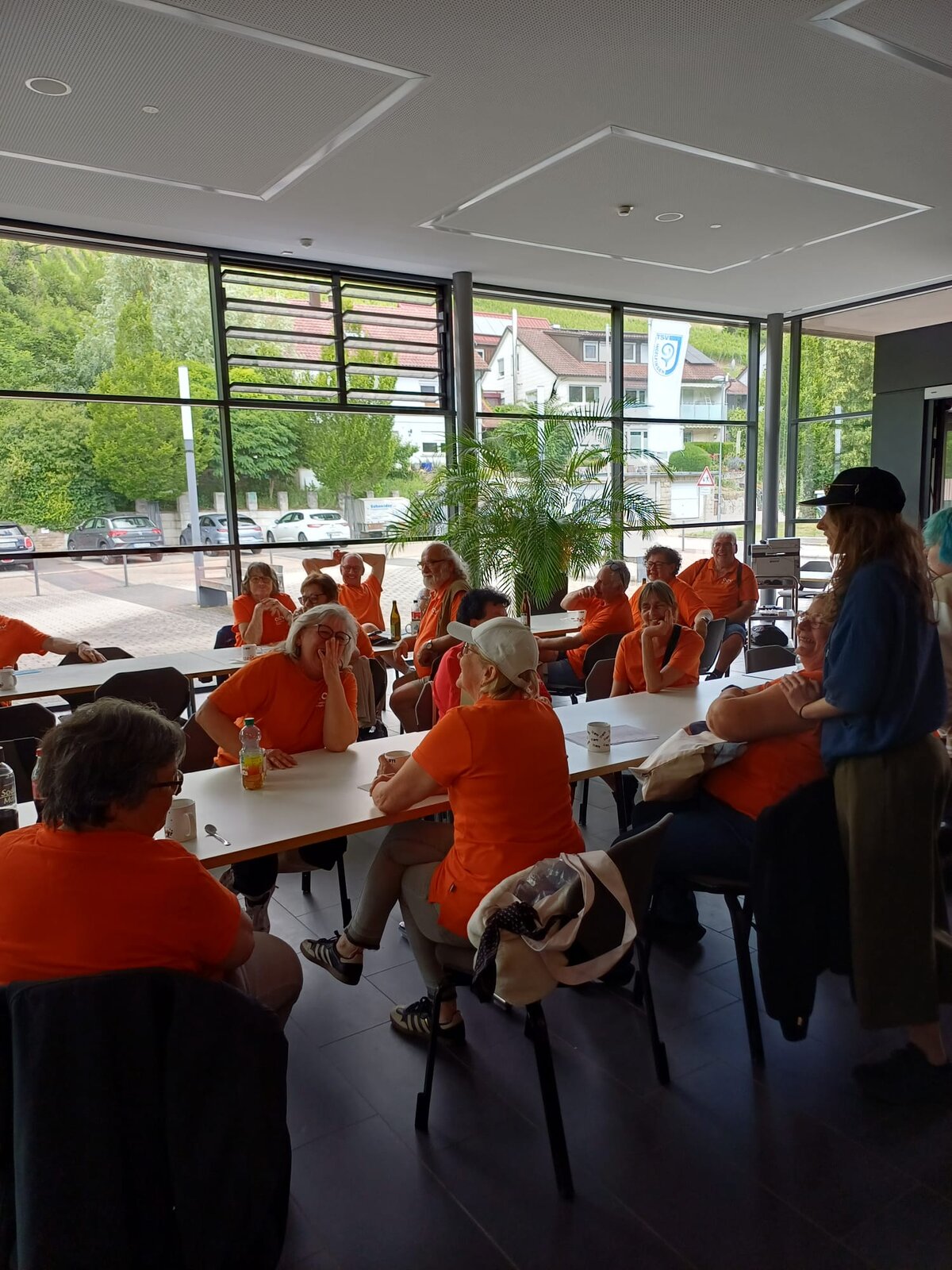 Eine Gruppe von Menschen in orangefarbenen Hemden sitzt in einem hellen Raum mit großen Fenstern an Tischen und unterhält sich. Einige hören einer stehenden Frau aufmerksam zu. Draußen sind Bäume und geparkte Autos zu sehen.