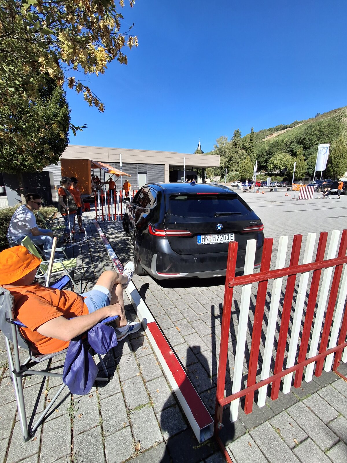 Ein schwarzes Auto fährt durch ein umzäuntes Gelände, während Menschen in orangefarbenen Hemden und Mützen an einem sonnigen Tag mit klarem Himmel und Bäumen im Hintergrund in der Nähe sitzen.
