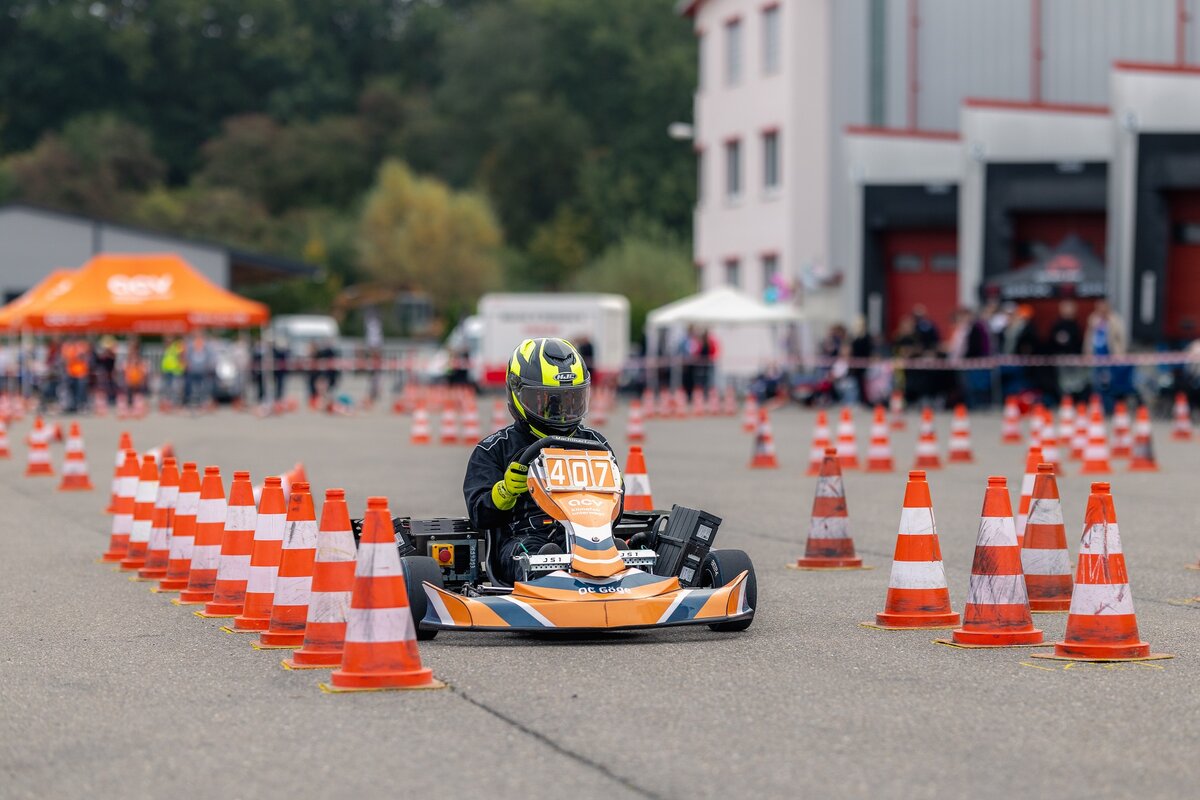 Ein Fahrer mit Helm und Rennanzug manövriert ein orange-schwarzes Go-Kart durch einen mit orangefarbenen Verkehrskegeln gesäumten Parcours.