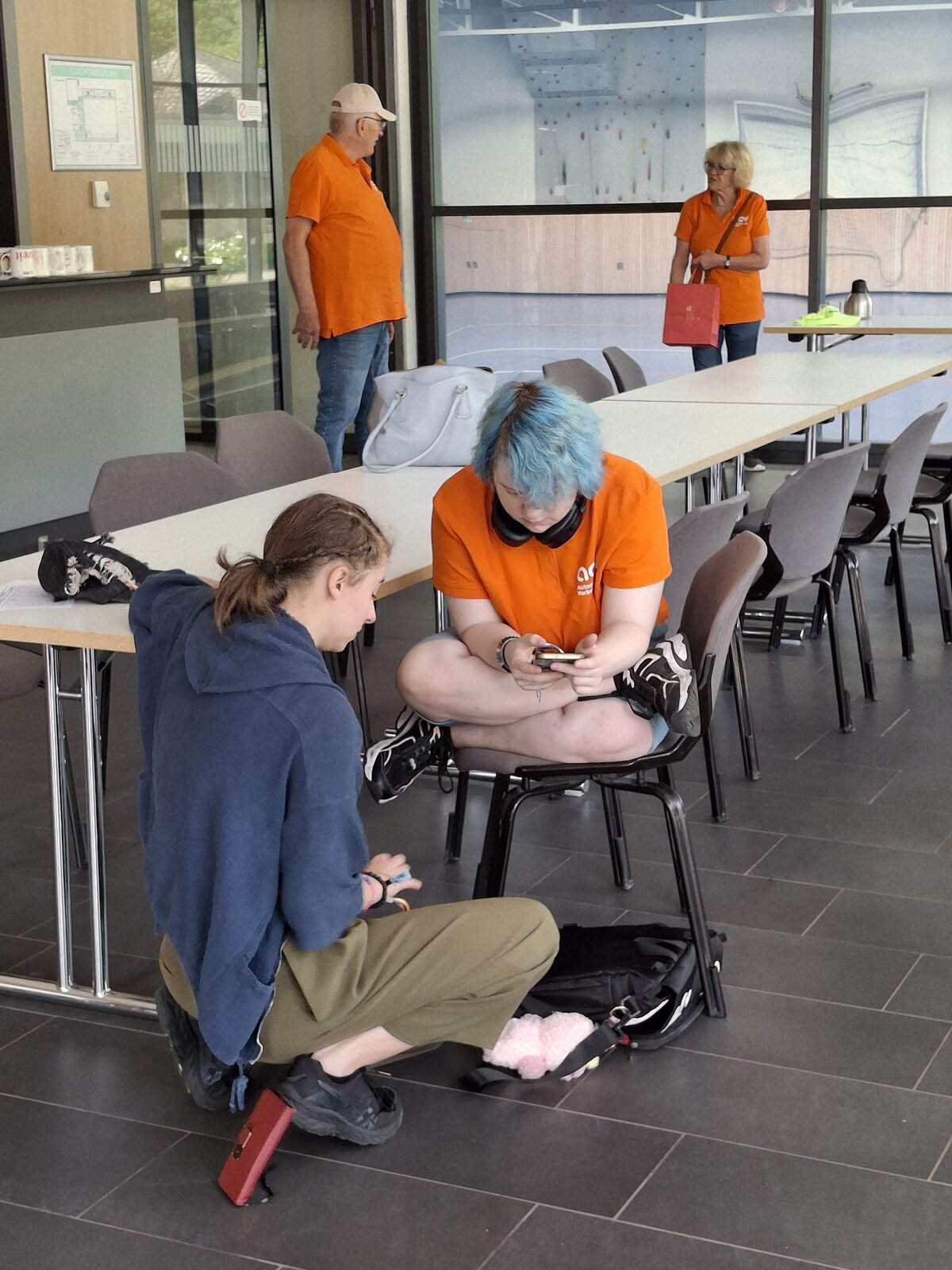 Zwei Personen sitzen dicht beieinander am Ende eines langen Tisches, eine auf dem Boden und eine mit gekreuzten Beinen auf einem Stuhl; beide sind auf ein Handy konzentriert. Zwei weitere Personen in orangefarbenen Hemden stehen im Hintergrund eines hellen Raums und unterhalten sich.