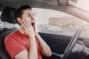 Junger Mann mit rotem T-Shirt sitzt schockiert am Steuer eines Autos, hält beide Hände an sein Gesicht, schaut mit weit geöffnetem Mund nach vorne – Szene vermittelt Panik im Straßenverkehr.