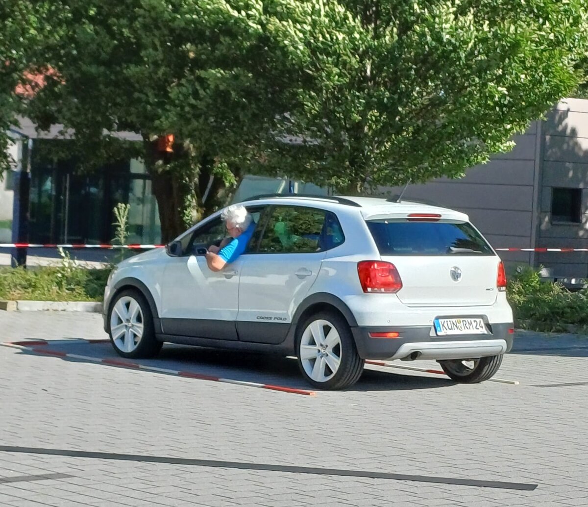 Eine Person mit grauen Haaren lehnt sich aus dem Fahrerfenster eines geparkten weißen Volkswagen Cross Polo, im Hintergrund sind Bäume und ein graues Gebäude zu sehen. Hinter dem Auto ist rot-weißes Absperrband zu erkennen.