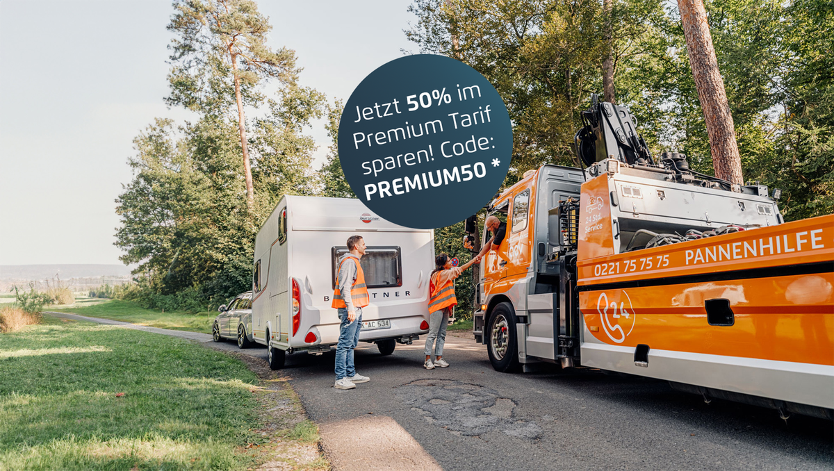 Ein großer orangefarbener Abschleppwagen hilft dabei, ein Wohnmobil am Straßenrand abzuschleppen. Zwei Personen in Warnwesten stehen neben dem Wohnmobil und dem Abschleppfahrzeug. Die Szene spielt auf einer Landstraße, die von hohen Bäumen gesäumt ist.