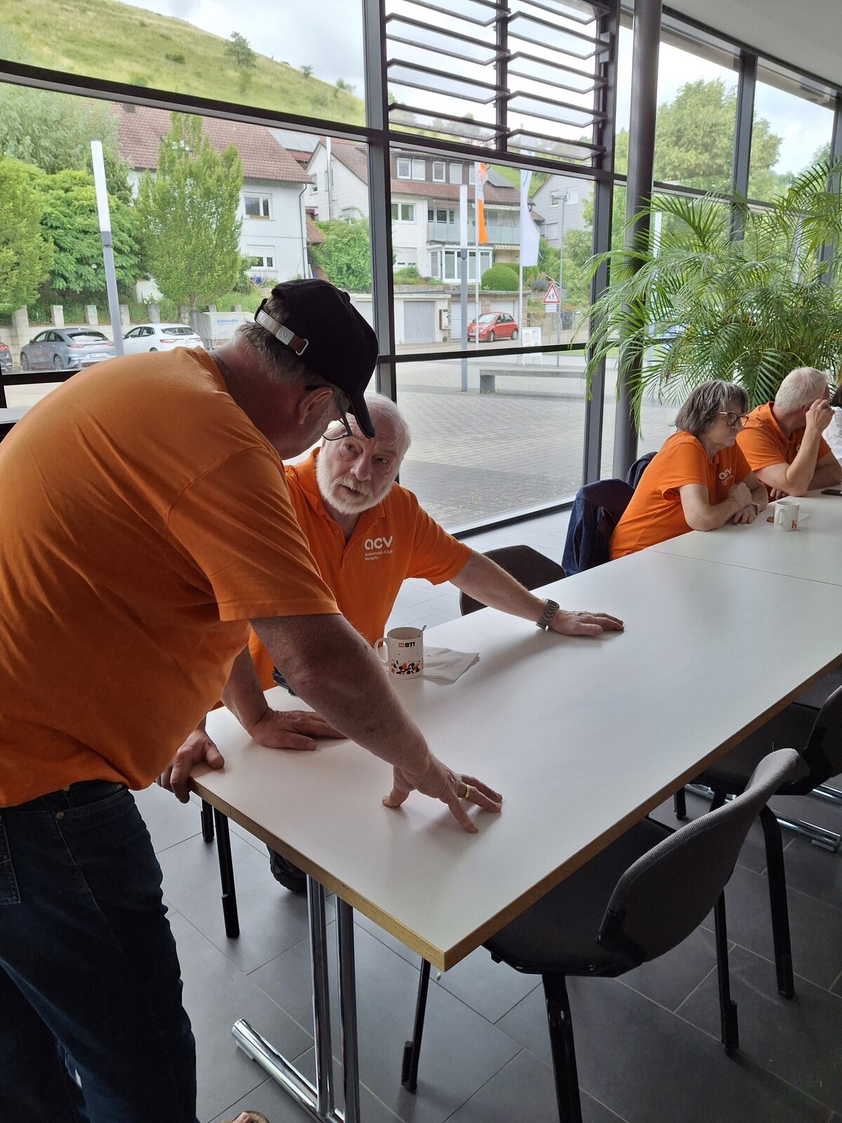 Zwei Männer in orangefarbenen Hemden unterhalten sich an einem weißen Tisch in der Nähe großer Fenster, während zwei weitere Personen, ebenfalls in orangefarbenen Hemden, mit Tassen daneben sitzen. Vor dem Fenster sind eine grüne Pflanze und Gebäude zu sehen.