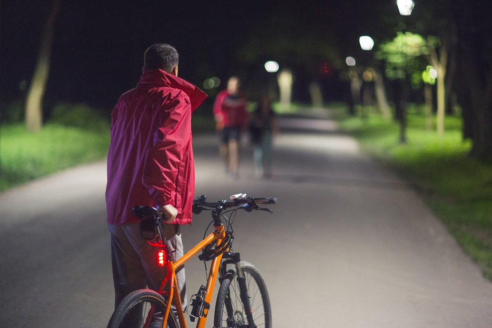 Ein Mann schiebt nachts sein Fahrrad auf einem beleuchteten Parkweg, im Hintergrund gehen Fußgänger auf ihn zu.