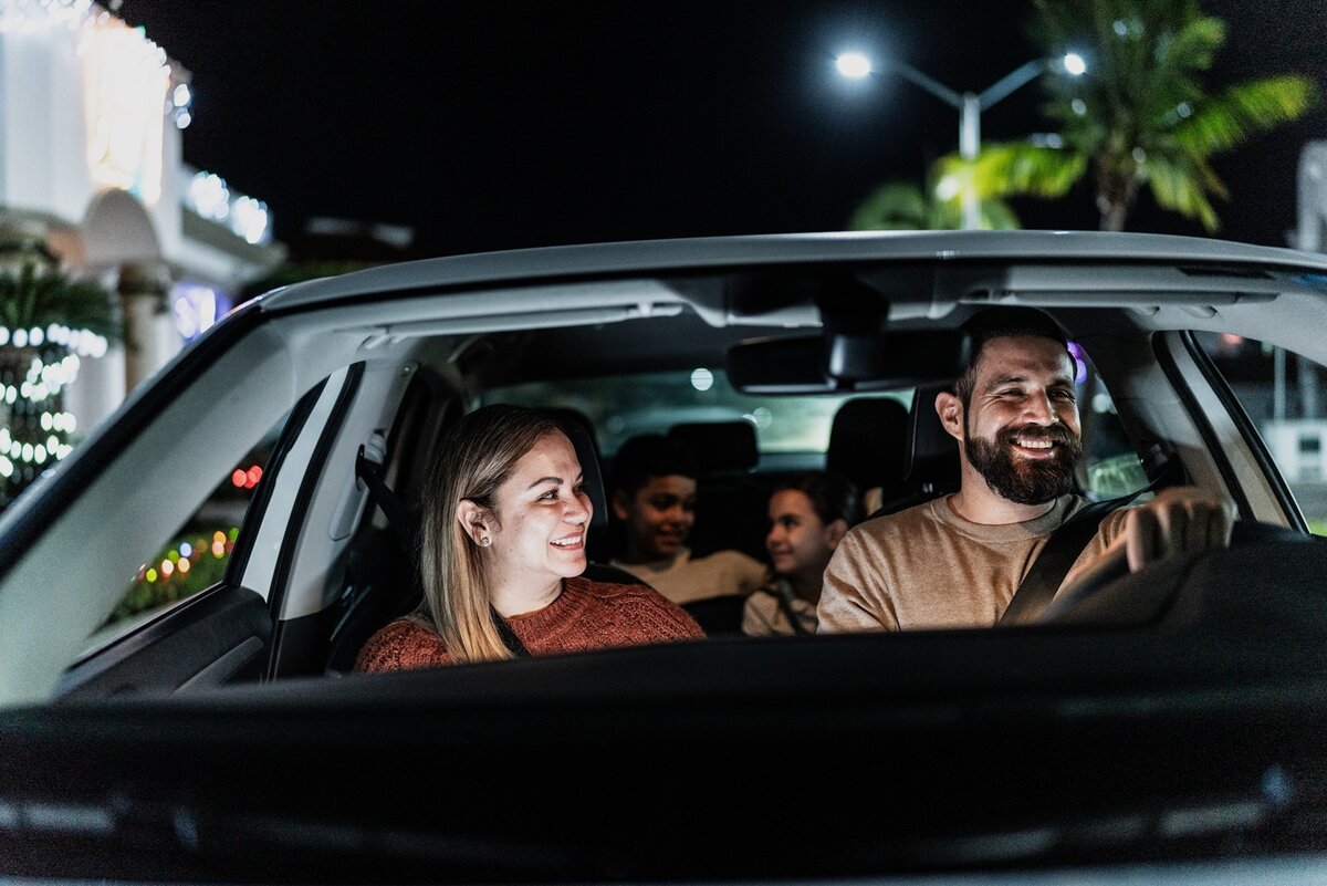 Zwei Erwachsene sitzen nachts lächelnd und plaudernd auf den Vordersitzen eines Autos, während zwei Kinder auf dem Rücksitz sitzen. Durch die Fenster sind die Lichter der Stadt und Palmen zu sehen.