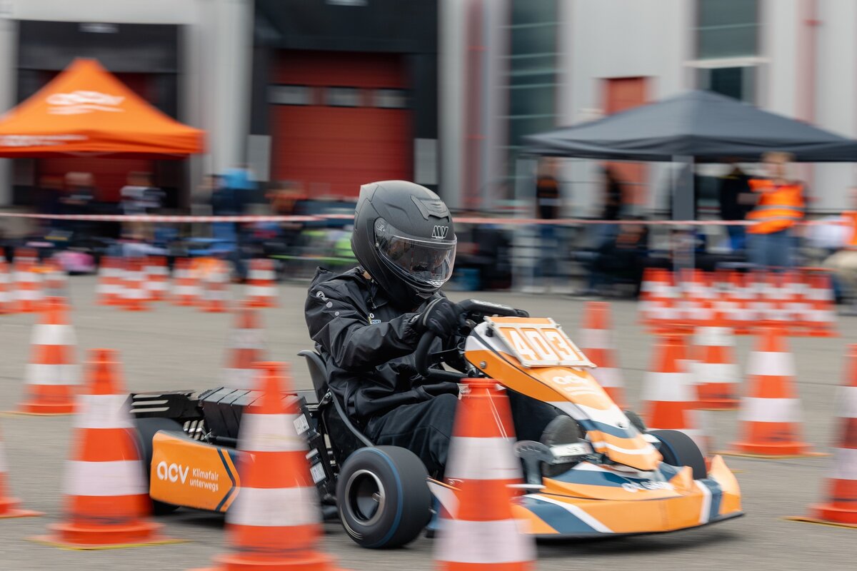 Eine Person mit schwarzem Helm und schwarzer Jacke fährt ein orange-schwarzes Go-Kart mit der Nummer 403 durch einen mit orangefarbenen Verkehrskegeln markierten Parcours.
