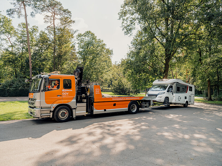 „Orangefarbener ACV Abschleppwagen ist vor einem weißen Wohnmobil positioniert um es abzuschleppen, beide Fahrzeuge stehen am Waldrand.“