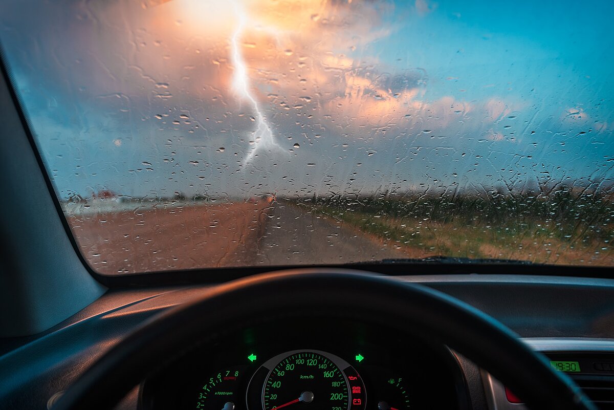 Blick aus dem Auto auf eine regennasse Straße mit Blitzeinschlag am Himmel – Gefahrensituation bei Gewitter.