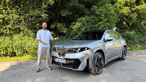 Ein Mann steht auf einem Feldweg neben einem silbernen BMW-SUV und deutet mit beiden Händen auf das Auto. Im Hintergrund sind an einem sonnigen Tag grüne Bäume und Büsche zu sehen.