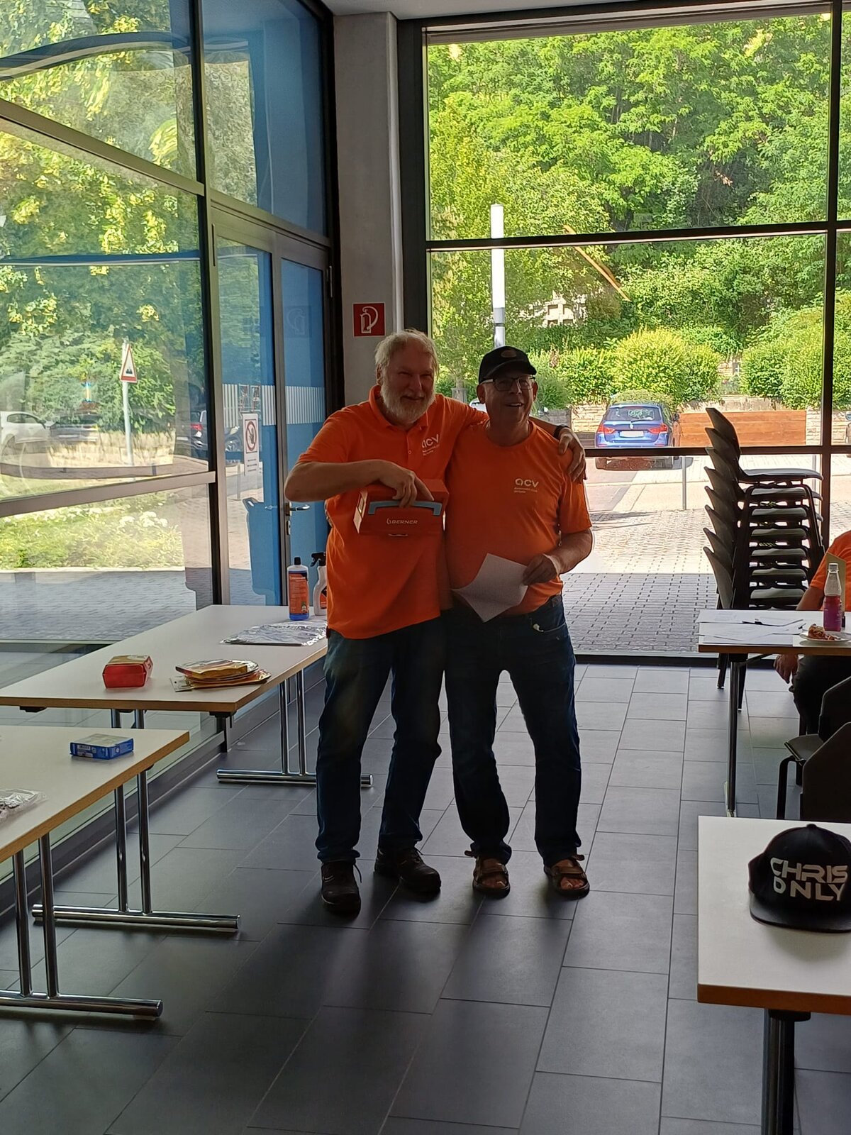 Zwei Männer in orangefarbenen Hemden stehen nebeneinander und lächeln in einem hellen Raum mit großen Fenstern. Einer von ihnen hält eine Kiste in der Hand. In der Nähe stehen Tische und Stühle; draußen sind Pflanzen und ein geparktes Auto zu sehen.