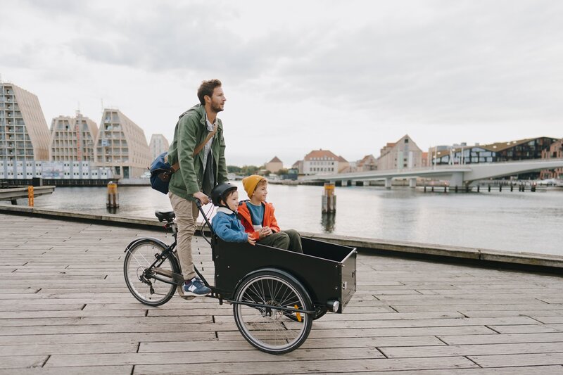 Ein Mann fährt mit einem Lastenrad, auf dessen Frontkiste zwei Kinder sitzen, auf einer Holzpromenade am Wasser entlang, im Hintergrund sind Stadtgebäude und eine Brücke zu sehen.