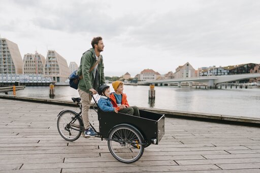 Ein Mann fährt mit einem Lastenrad, auf dessen Frontkiste zwei Kinder sitzen, auf einer Holzpromenade am Wasser entlang, im Hintergrund sind Stadtgebäude und eine Brücke zu sehen.
