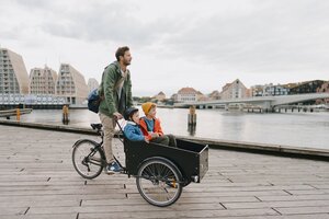 Ein Mann fährt mit einem Lastenrad, auf dessen Frontkiste zwei Kinder sitzen, auf einer Holzpromenade am Wasser entlang, im Hintergrund sind Stadtgebäude und eine Brücke zu sehen.