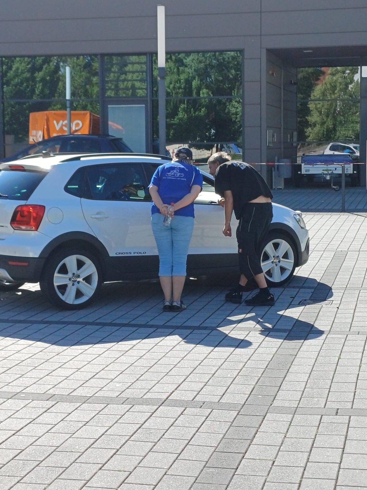 Zwei Personen stehen neben einem weißen Volkswagen Cross Polo. Eine Person in blauem Hemd und Jeans hält eine Flasche in der Hand. Die andere Person, die ein schwarzes T-Shirt und eine Jogginghose trägt, beugt sich über das Auto.