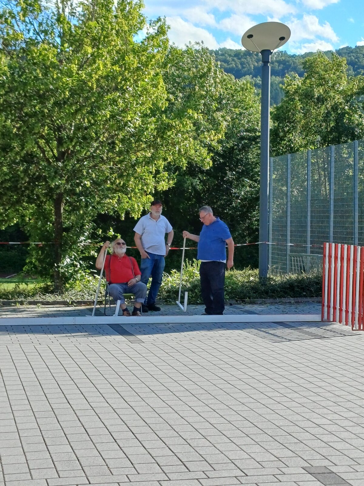 An einem sonnigen Tag haben sich drei ältere Menschen im Freien versammelt; eine Frau sitzt, während zwei Männer neben ihr stehen und sich unterhalten. Sie befinden sich in der Nähe von Bäumen, einem Drahtzaun und einer gepflasterten Fläche. Außerdem sind ein Laternenpfahl und eine rot-weiße Absperrung zu sehen.