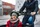 Das Bild zeigt ein fröhliches Kind, das in einer Transportbox eines Lastenfahrrads sitzt. Das Kind trägt eine rote Jacke, einen grünen Helm und einen dicken Schal, passend zur kühlen Jahreszeit. Hinter dem Kind ist ein Erwachsener in dunkler Kleidung zu sehen, der das Fahrrad lenkt. Der Hintergrund zeigt eine städtische Umgebung mit Gebäuden und Straßen, was auf eine urbane Fahrradtour hinweist. Die Szene vermittelt eine warme und sichere Atmosphäre.